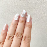 Close - up of fingers wearing ethereal floral embellished press - on nails with a soft pearl finish against a light background