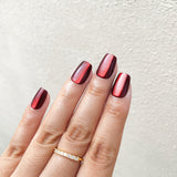 Close - up of fingers wearing ruby red holographic square press - on nails with a high - shine finish, accompanied by a gold diamond - encrusted ring against a light background