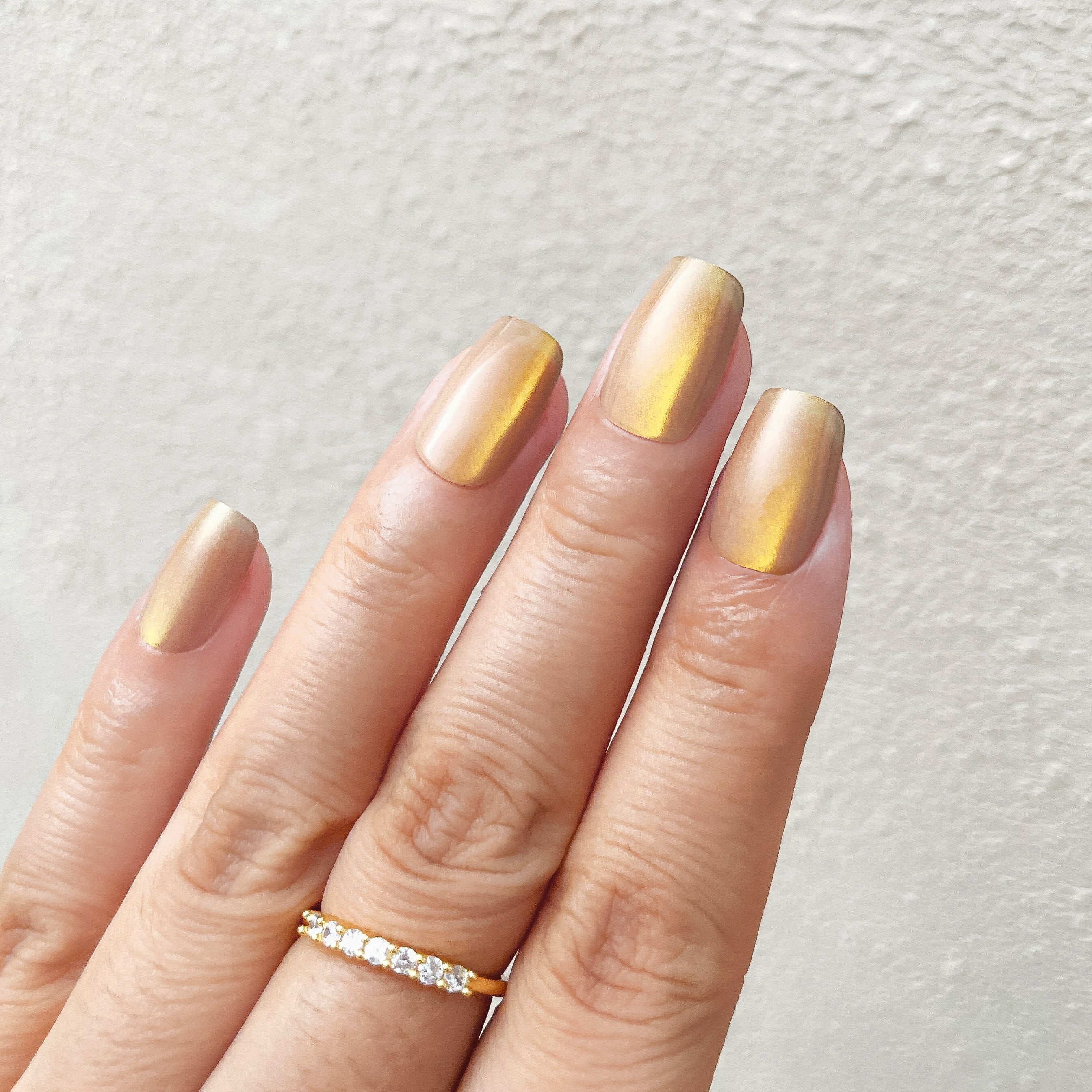 Close - up of fingers wearing gold - brown gradient metallic press - on nails, paired with a delicate diamond - studded band against a light background