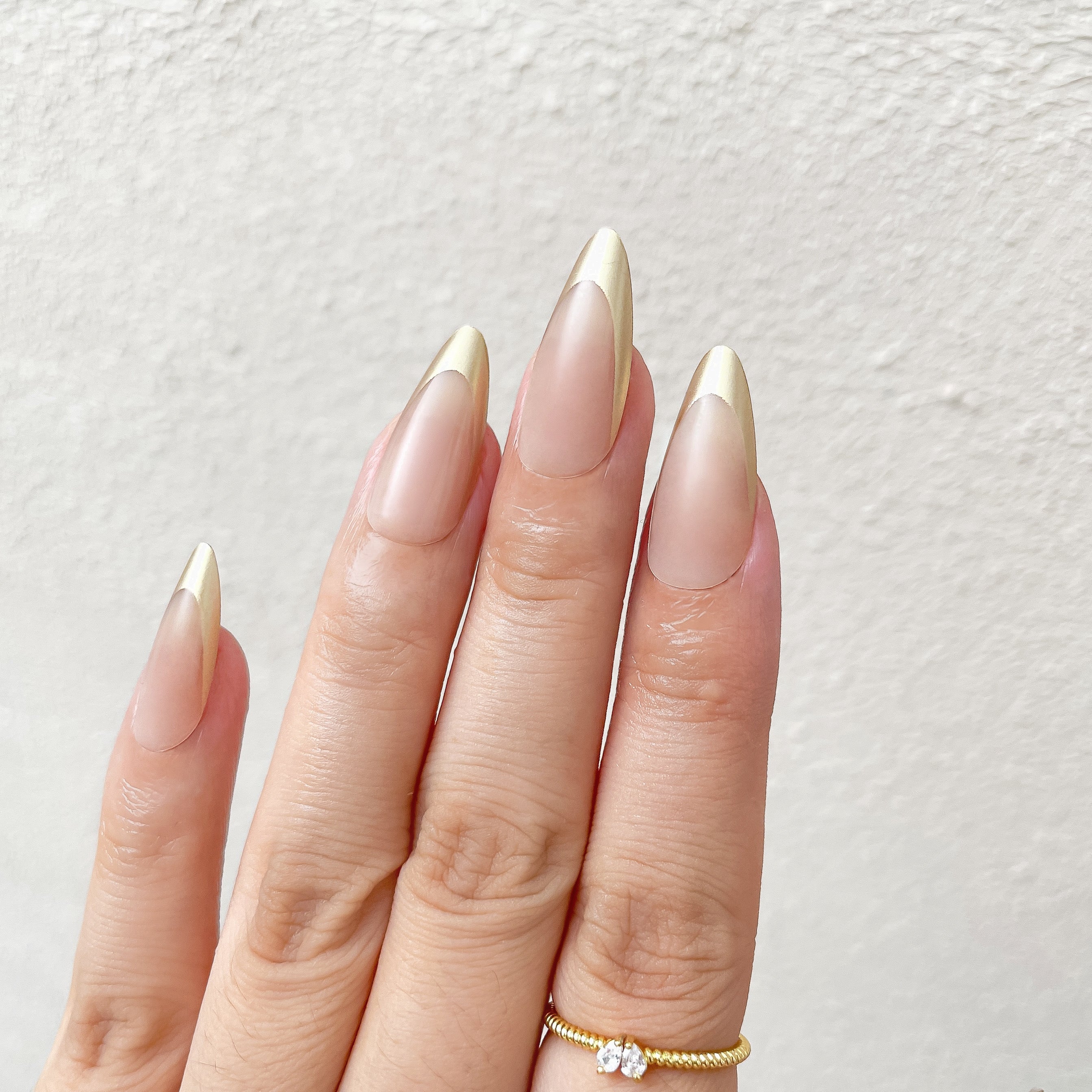 Hand with long, pointed nails painted in a gradient of beige and gold on a light background.